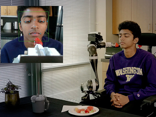 Robotic arm feeding seated person a strawberry on a fork with an inset image of the robot mapping features of their face