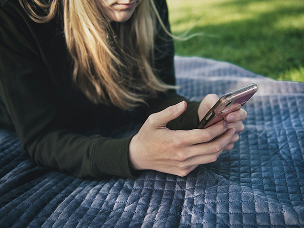 A person with long blond hair, with only mouth and chin visible, is lying on a blue quilted blanket on short green grass in dappled sunlight. The person is wearing a black sweatshirt and propped up on their elbows, viewing a smartphone held in their well-manicured hands.