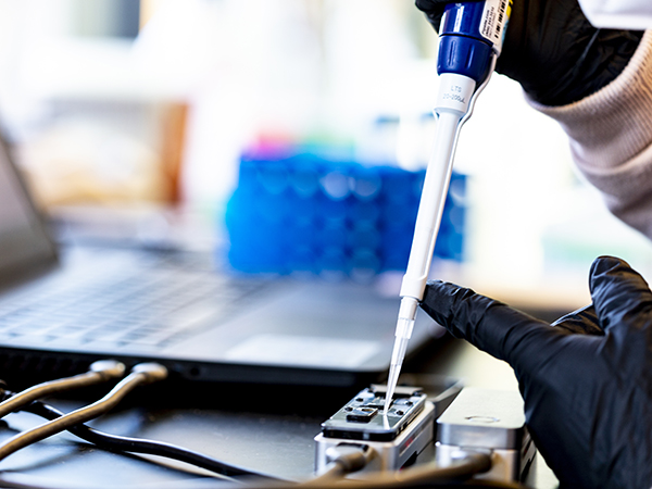 Gloved hands piping liquid into a smalll rectangular nanopore device connected to a laptop