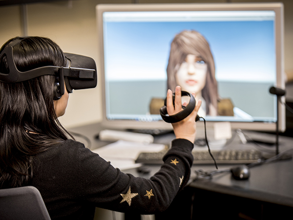 A student wearing an Oculus headset motions toward a screen displaying a virtual character