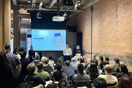 A brick lined room full of people watching a presentation by a student in front of a PowerPoint screen displaying the company name Stimma