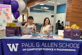 Two people seated at a table draped with an Allen School tablecloth and displaying swag and balloons make number-one signs with their fingers