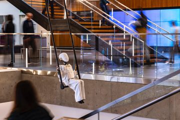 A robotic acrobat that resembles the figure of a boy sits on a trapeze suspended above a stairwell in the Gates Center atrium as people walk past