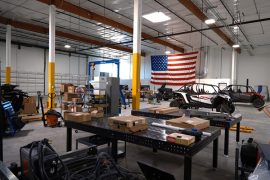 A factory floor with tables and shelves containing boxes interspersed with machinery and vehicle parts