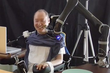 A person in a wheelchair is seated at a table behind a mouth-controlled laptop, with a robotic arm alongside moving a forkful of food towards his mouth as the person smiles